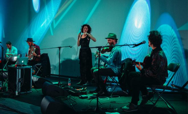 Four musicians playing in a concert. A man playing drums, a man playing accordion, a woman singing, a man playing violin and a man playing guitar