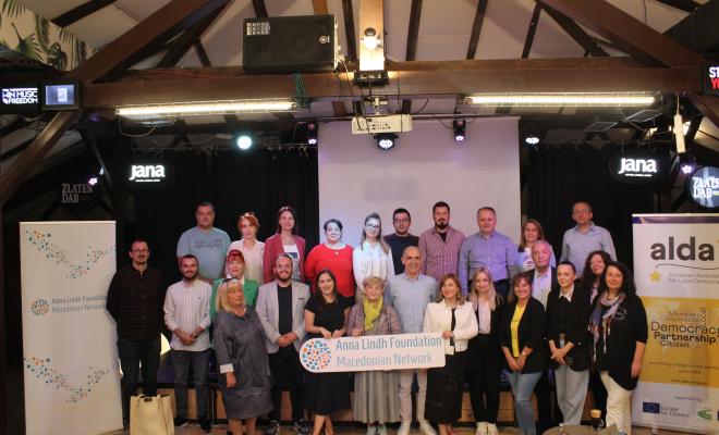 Group photo of members of the Macedonian Network of the Anna Lindh Foundation, taken at the 'Building Connections, Creating Opportunities' event in Public Room. Representatives from various organizations stand together, holding a sign with the network's name, surrounded by banners of ALDA and the Anna Lindh Foundation