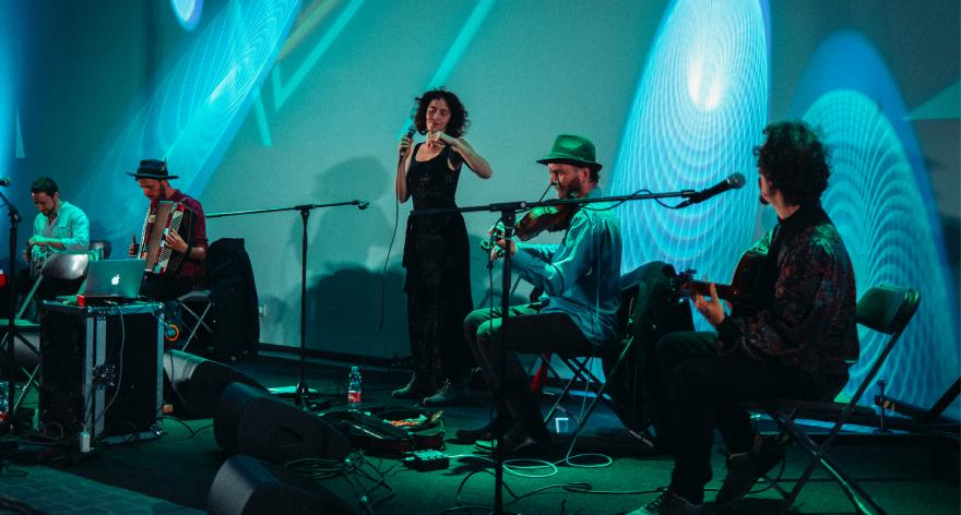 Four musicians playing in a concert. A man playing drums, a man playing accordion, a woman singing, a man playing violin and a man playing guitar